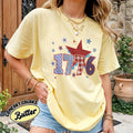 Person wearing a yellow t-shirt with '1776' design, standing outdoors.
