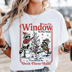 A woman wearing a white graphic t-shirt with a Christmas-themed raccoon design that reads 'From The Window To The Walls' and 'I'm About To Deck These Halls'.