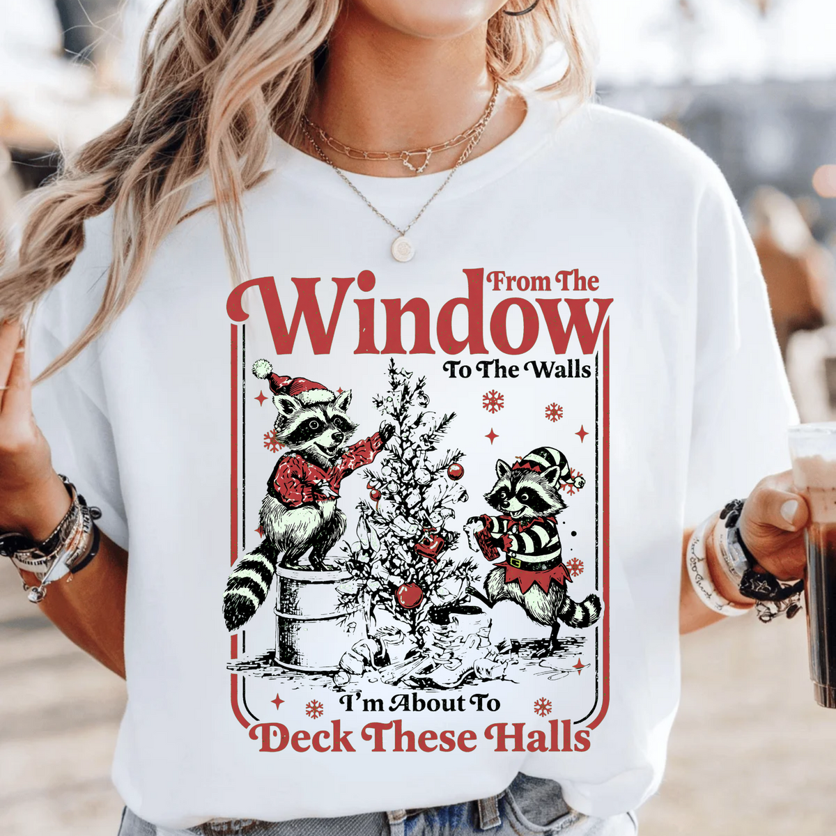 A woman wearing a white graphic t-shirt with a Christmas-themed raccoon design that reads 'From The Window To The Walls' and 'I'm About To Deck These Halls'.