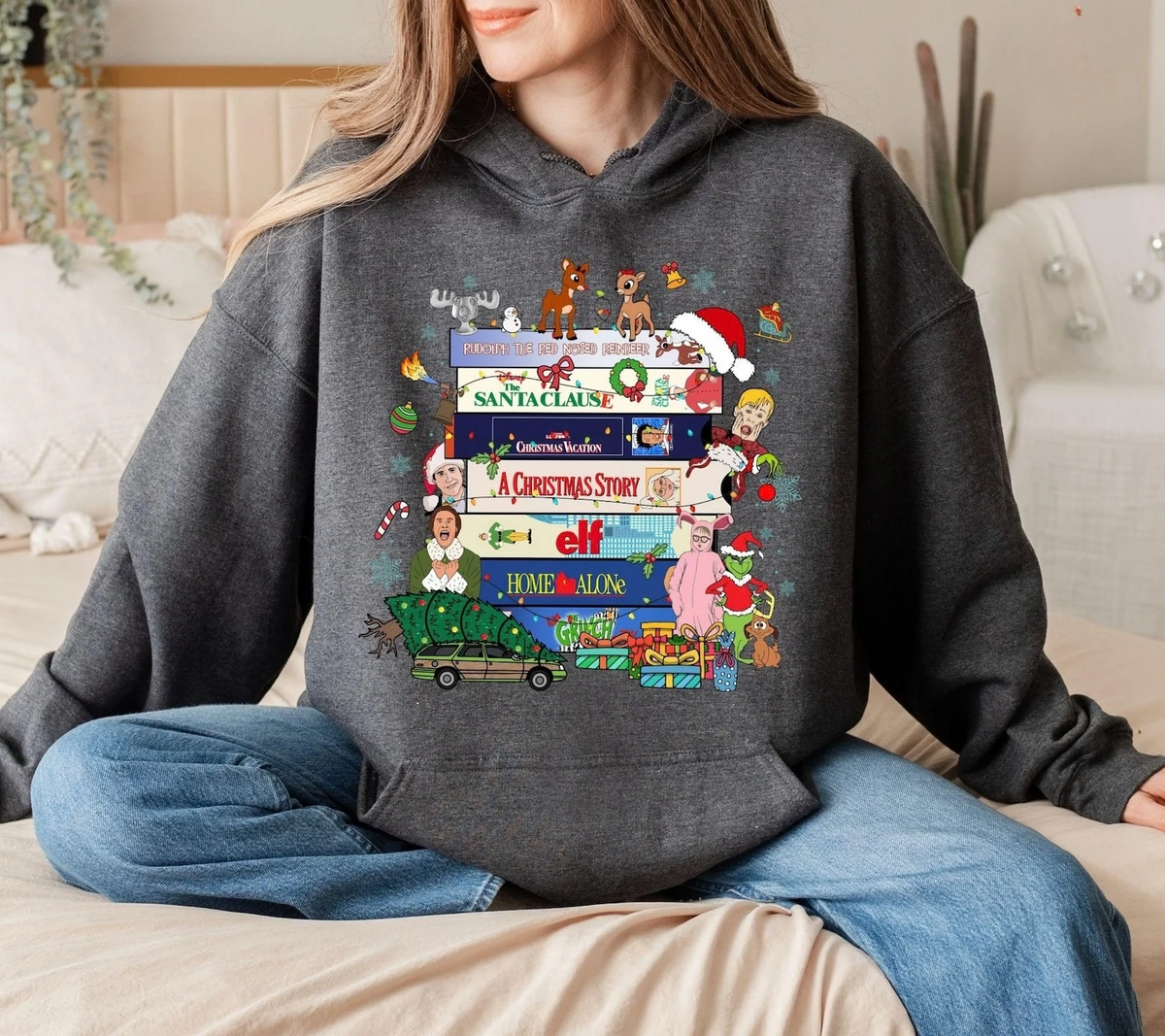 A woman wearing a dark gray hoodie with a colorful Christmas-themed graphic print featuring movie quotes and characters, sitting on a bed in a cozy bedroom setting.