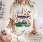 A woman wearing a cream-colored t-shirt with a colorful Christmas-themed graphic print featuring Rudolph the Red-Nosed Reindeer, Santa Claus, and holiday books, sitting on a bed and holding a white mug.