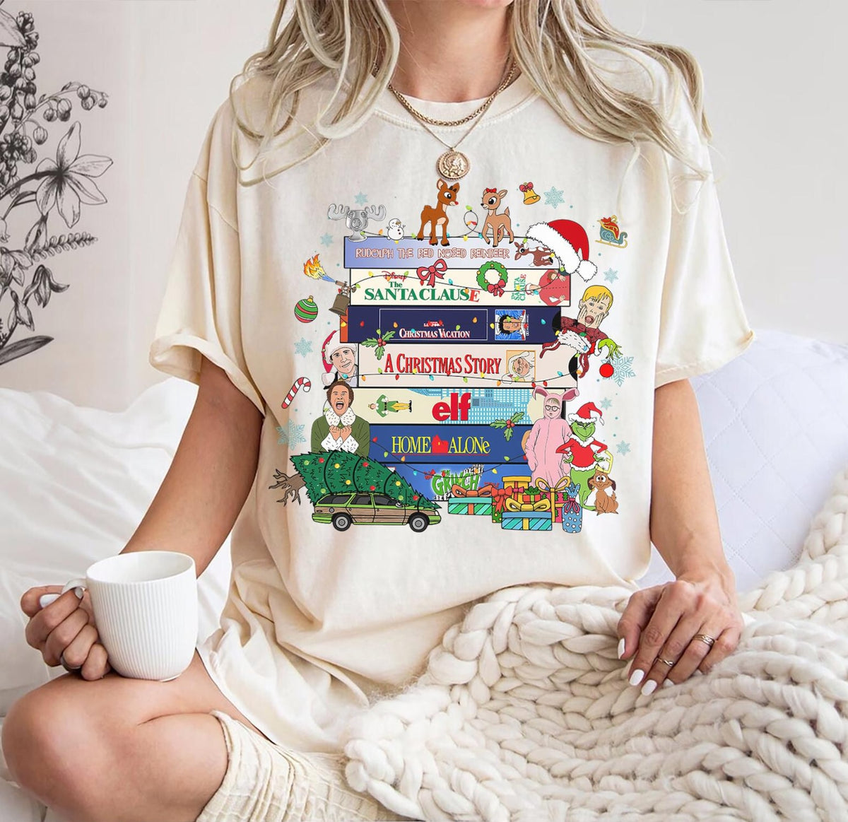 A woman wearing a cream-colored t-shirt with a colorful Christmas-themed graphic print featuring Rudolph the Red-Nosed Reindeer, Santa Claus, and holiday books, sitting on a bed and holding a white mug.