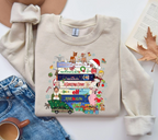 A beige crewneck sweatshirt laid flat on a white surface, featuring a colorful Christmas-themed graphic print with movie quotes and characters. The sweatshirt is styled in a flat lay with blue jeans, a coffee cup, an open book, and a dry autumn leaf.