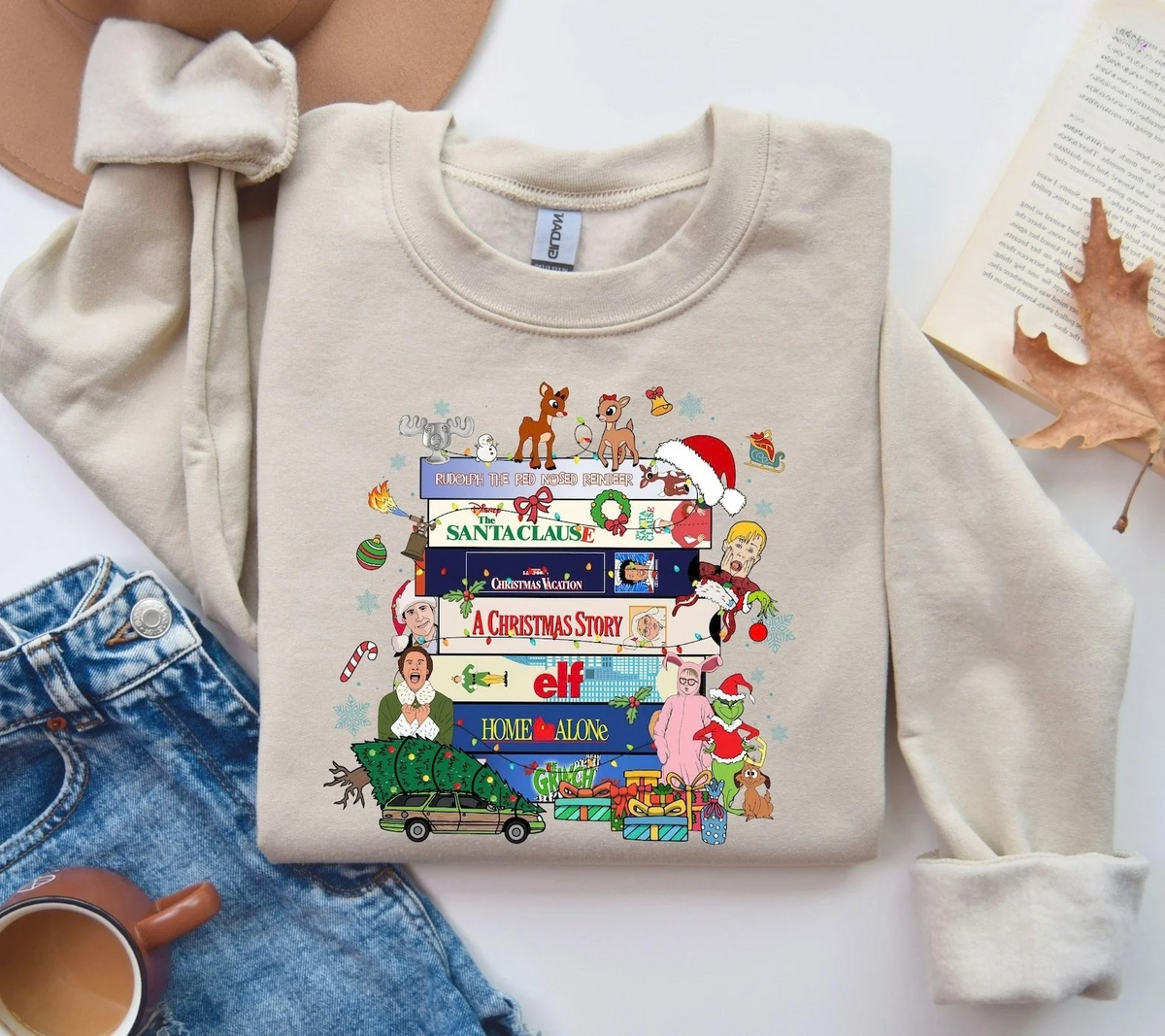 A beige crewneck sweatshirt laid flat on a white surface, featuring a colorful Christmas-themed graphic print with movie quotes and characters. The sweatshirt is styled in a flat lay with blue jeans, a coffee cup, an open book, and a dry autumn leaf.