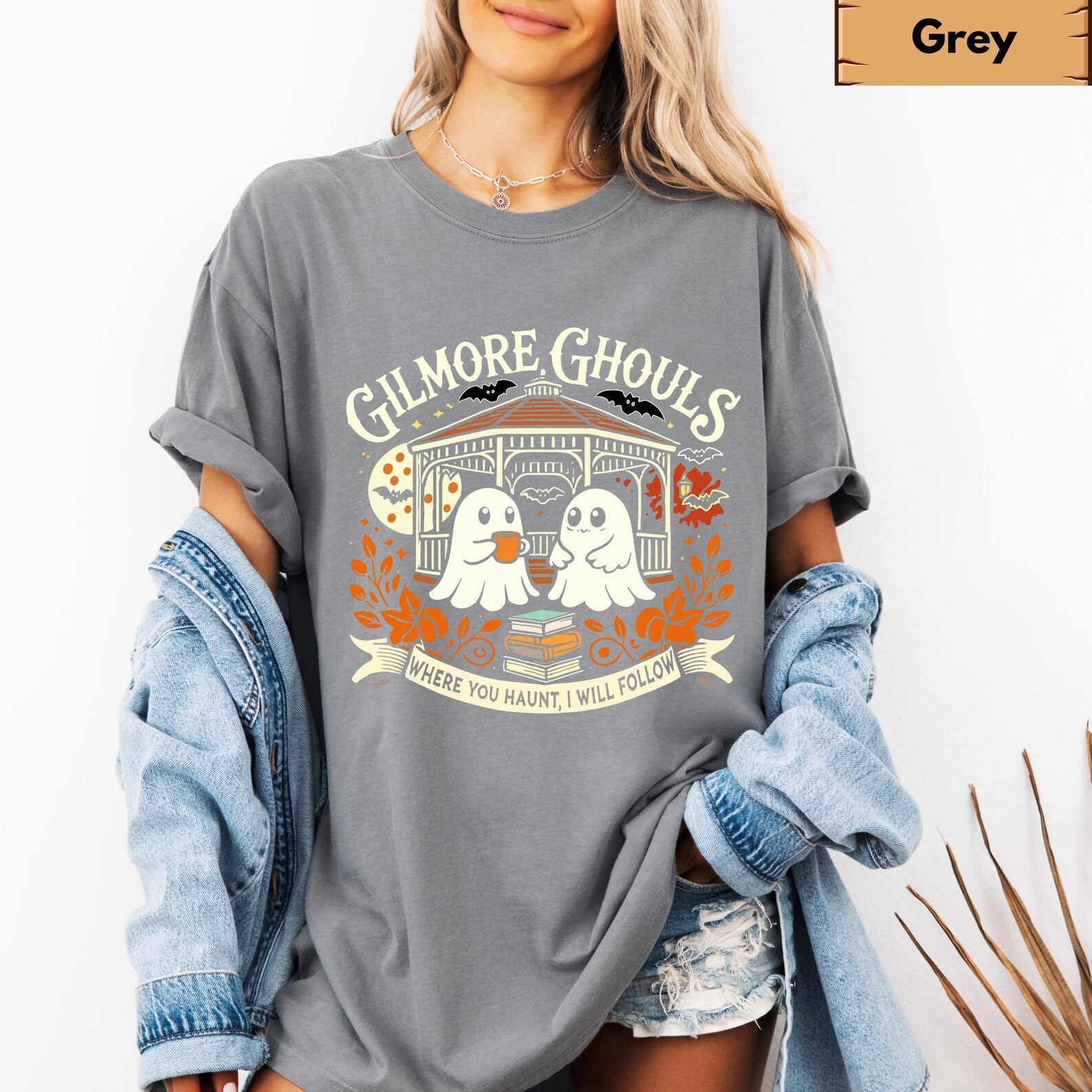 A woman wearing an oversized heather gray graphic t-shirt with a 'Ghimore Ghouls' Halloween design, styled with a light blue denim jacket draped off her shoulders and distressed denim shorts.